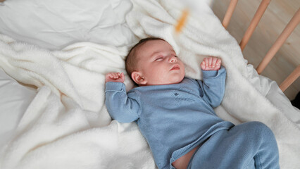 Baby boy sleeping with clenched fists and curled legs in a blue knit outfit on a white fleece blanket inside a wooden crib  gentle serenity. © Krakenimages.com