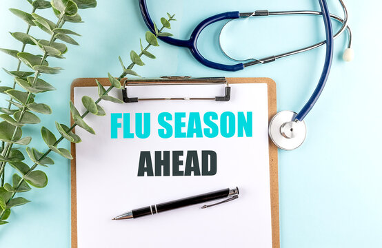 Flu season ahead text on a clipboard with stethoscope, pen and green leaves on a blue background, influenza prevention
