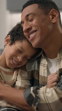 Smiling African American father using mobile phone scrolling social media while little boy son child hugging parent use smartphone online happy family dad preschooler kid indoor home living room couch