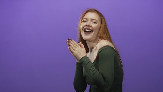 Redhead woman claps hands showing exposed shoulder and smile against purple studio backdrop, long hair falling down her back; joyful warmth.