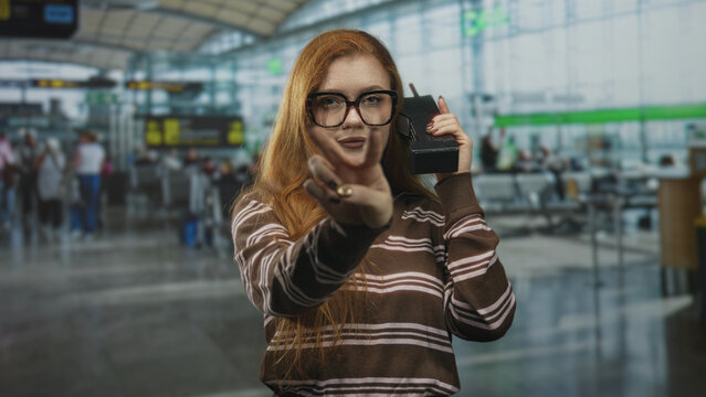 Redhead woman holding retro cellphone and showing v sign with her hand in a busy airport terminal; playful confidence.