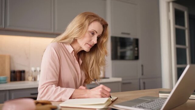 Mature Caucasian blonde woman writing notes in notebook while working with laptop at home. Focused female planning tasks and organizing schedule while sitting at table in modern kitchen.