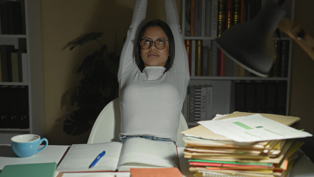 Young hispanic woman studying at night, writing in a notebook then leaning back and stretching arms with eyes closed, desk piled with folders, pen and mug by a lamp in studio; fatigue perseverance.