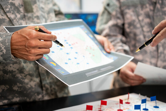 Military personnel in camouflage uniforms review a printed map and tactical markers during a planning briefing.