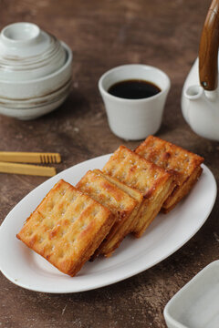 Traditional Indonesian gabin tape crackers filled with sweet fermented cassava, golden brown crispy biscuits served on a white ceramic plate with black coffee and a teapot on a rus