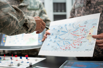 Fototapeta premium Military personnel in camouflage uniforms review a tactical map during a planning meeting, with markers, documents and a digital screen on the table.