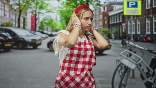 Woman cook in apron covers ears with hands on street near parked cars and bicycles; stress noise overload.