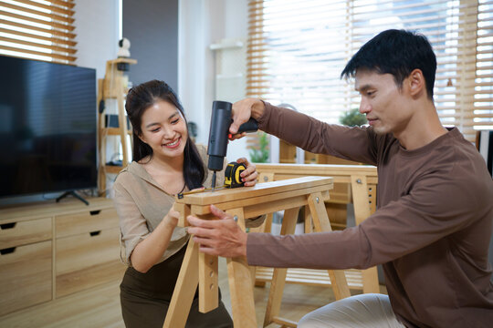 Happy Asian couple collaborating to assemble flat pack wooden furniture on living room floor, ideal for home improvement and DIY lifestyle concepts