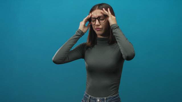Woman wearing glasses presses her temples and winces, showing a headache while standing against a blue studio wall; stress fatigue.