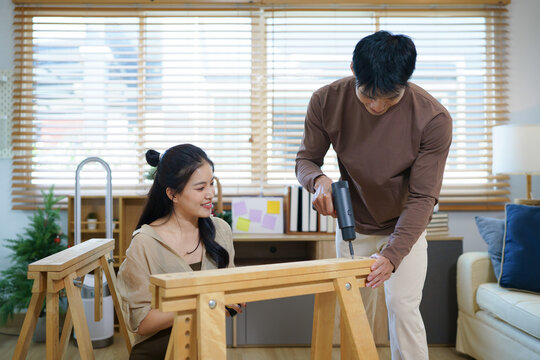 Happy Asian couple collaborating to assemble flat pack wooden furniture on living room floor, ideal for home improvement and DIY lifestyle concepts