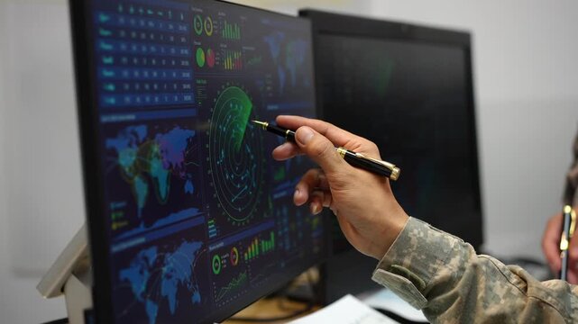 Close-up of a person in camouflage pointing with a pen at digital dashboards showing maps, radar graphics, charts and security data on computer screens