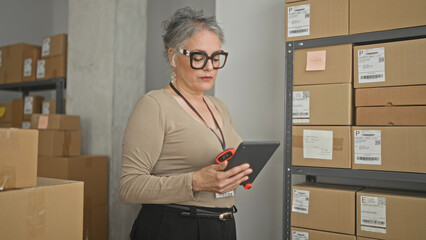 Woman holding tablet and red scanner scans stacked cardboard shipping boxes on metal shelving in a small storage building  focused efficiency. © Krakenimages.com