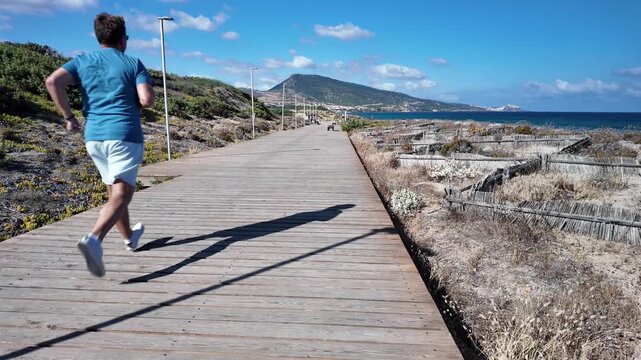 A middle-aged man is running along a wooden boardwalk by the seashore, with dunes and hills visible in the background.