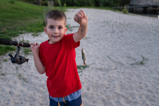 A 6-year-old boy with a fishing rod shows off a caught fish against the backdrop of a pond or river; active leisure.