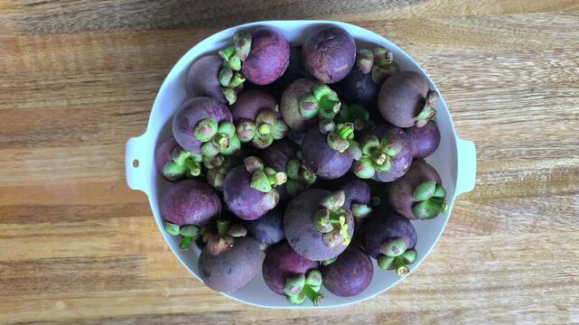 A white plastic bowl filled with fresh mangosteen fruits. Placed on a wooden table, it highlights the rich dark purple skin and vibrant natural texture of the tropical fruit.