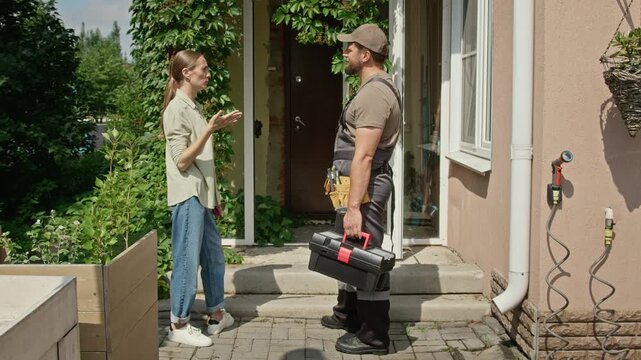 Caucasian young adult woman explaining repair task to Caucasian craftsman holding toolbox at house entrance. Clip suits home improvement campaigns, renovation services and maintenance promos