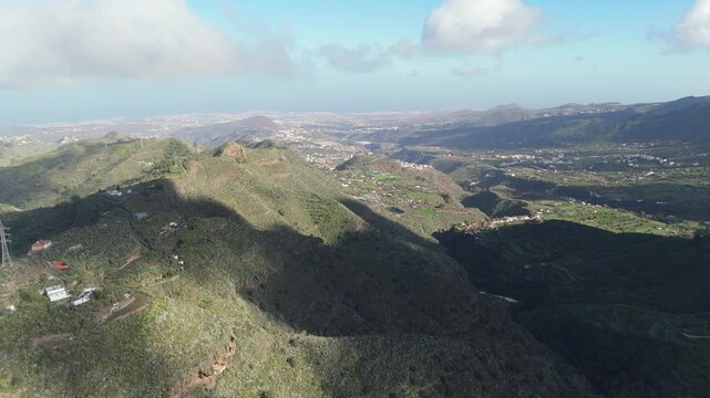 Aerial drone push in shot of the rugged, green landscape around Monta&ntilde;a de los Cardos and El Monta&ntilde;&oacute;n on Gran Canaria, revealing distant villages and the ocean.