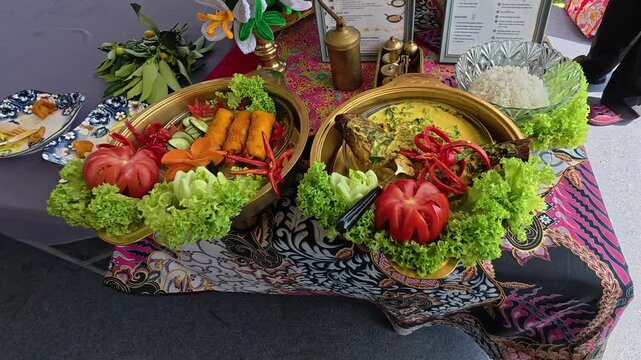 A professional chef plates and garnishes a modern Malay fusion dish during a cooking competition. The beautifully decorated meal is presented on a table for judges to evaluate.