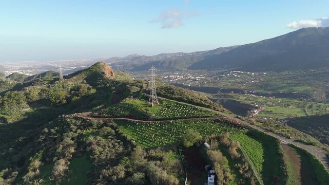 Aerial drone push in shot revealing the rugged, lush landscape of Monta&ntilde;a de los Cardos on Gran Canaria, Canary Islands. Captures green hills, distant villages, and the ocean.