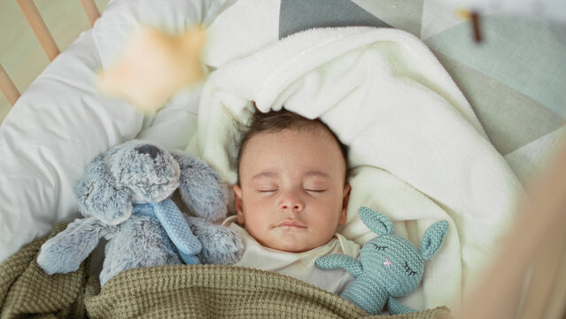 Baby boy sleeping in a cradle with plush bunny and blanket, face and closed eyes visible in a building; serenity comfort.