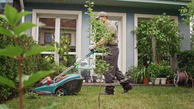 Caucasian young adult gardener mowing backyard lawn using electric mower outside modern house. Useful for landscaping company ads, gardening equipment promos, or home care videos