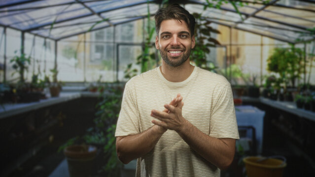 Man claps hands in a greenhouse building among potted plants and terracotta pots, smiling and rubbing his palms together; quiet joy.