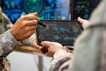 Military personnel in camouflage use a stylus and tablet to review maps, charts, and digital information in a modern command center. © Thitaporn