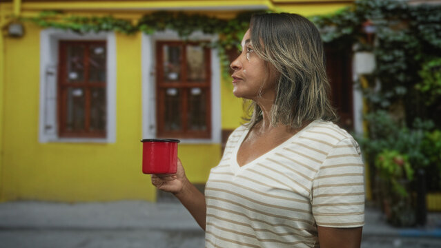 Woman holding red cup at chest level on a colorful street in front of a yellow building with window and plants; quiet contemplation.