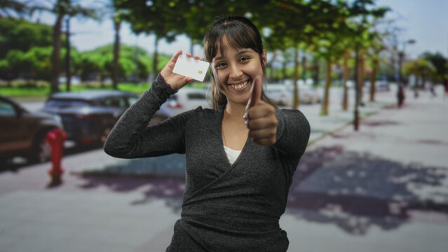 Woman holding creditcard showing thumb down gesture on a street with parked cars and trees; consumer distrust disapproval.