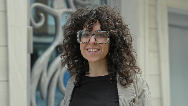 Woman wearing oversized tortoiseshell glasses smiles at camera showing teeth while standing by decorative metal sculpture and stone facade on street; confidence warmth.