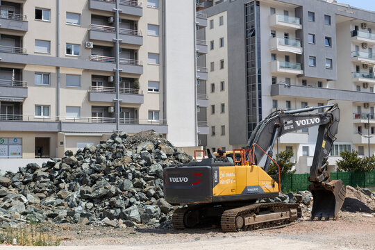 July 1, 2026, Budva, Montenegro, Excavator Working Near Residential Buildings with Pile of Rocks and Construction Materials on Site