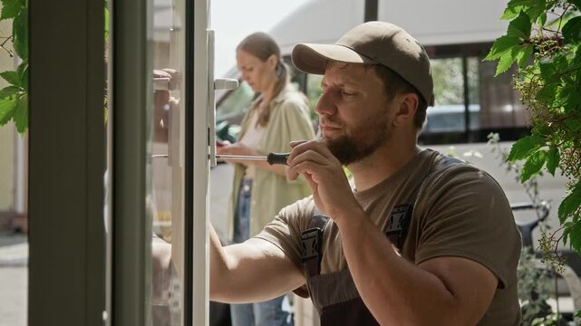 Caucasian handyman fixing glass door lock using screwdriver while female client stands in background. Visual fits locksmith service commercials, home improvement and repair advertising