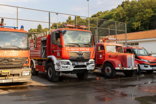 June 26, 2026, Petrovac, Montenegro, A lineup of modern and vintage fire trucks parked in a lot near a sports court and trees under a clear sky