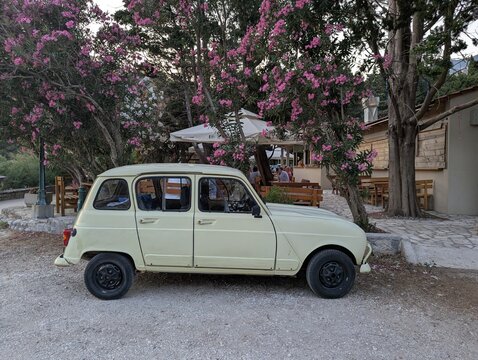 June 28, 2026, Petrovac, Montenegro, Vintage yellow car parked beside flowering trees near a casual outdoor restaurant with wooden seating