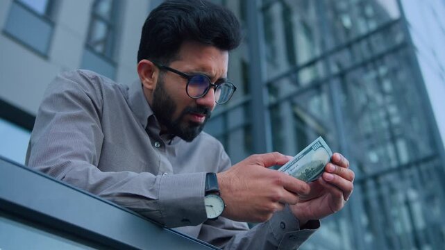 Worried Indian Arabian businessman counting dollar cash outdoors in city upset poor man male calculating salary income profit facing financial crisis miserable entrepreneur debt tight budget inflation