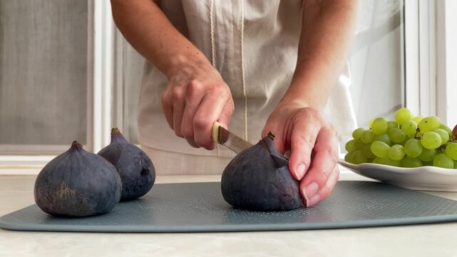 A woman is cutting figs on a gray cutting board. Close‑up of a woman&rsquo;s hands cutting the fruit with a knife, a simple and healthy snack