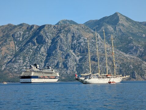 July 4, 2026, Kotor, Montenegro, A large cruise ship and a tall sailing ship anchored in a calm bay surrounded by mountains under a clear blue sky