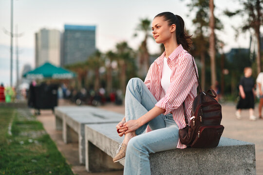 Woman lifestyle outdoor portrait seated on a concrete ledge with backpack beside, wearing striped shirt and jeans, relaxed mood and thoughtful gaze in a sunny city park during afternoon stroll under