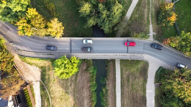 Aerial view of cars crossing a narrow bridge over a small stream beside rural homes and autumn trees in a green countryside landscape. Traffic at country landscape surrounded by trees, grassy fields, 