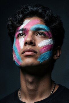 Young trans person with colorful pride flag paint on face. Artistic portrait shows confident gaze looking up. Male model creative makeup with blue pink white strokes. Expressive face art promotes