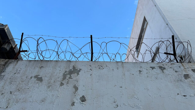 Rusty barbed wire on top of an old weathered white wall against a clear blue sky.