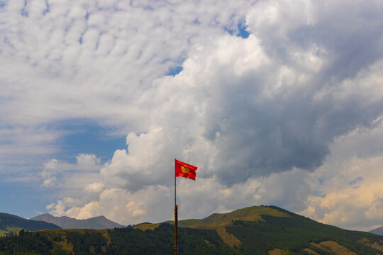 Red national flag of Kyrgyzstan waves from a pole against a backdrop of green mountains and hills. Partly cloudy sky adds dynamic weather to the natural outdoor scene.