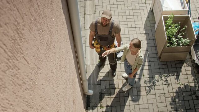 Young adult Caucasian woman showing exterior wall damage to Caucasian handyman holding toolbox from top view. Perfect for home repair marketing and contractor promotional campaigns
