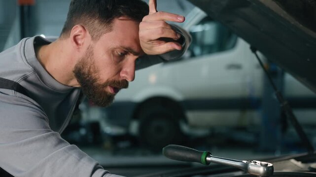 Exhausted Caucasian man mechanic wiping sweat while repairing engine in garage male technician using spanner on car motor tired worker fixing truck breakdown automotive service physical labor fatigue
