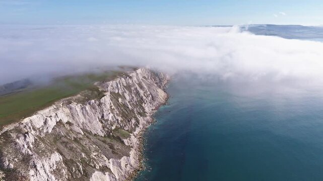 High aerial shot of white chalk sea cliffs meeting blue ocean water with a dense sea fog cloud inversion bank rolling over the green headland.