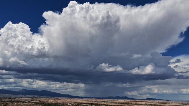 4K drone time lapse capturing a towering cumulonimbus thunderhead drifting across an arid southwestern plateau. A dark underbelly drops visible rain shafts onto the rugged terrain below.