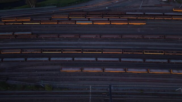 Top down aerial drone flight over a large railway yard in England, showing rows of colorful freight wagons and cargo cars arranged across multiple parallel tracks. Dark rails and orange, yellow, brown