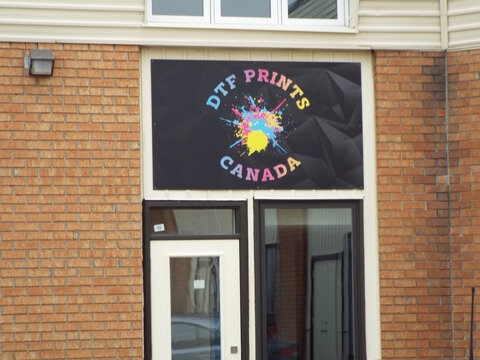Gloucester, Ontario, Canada &ndash; August 1, 2026: A colorful business sign for "DTF Prints Canada" with a paint splatter graphic is mounted on a brick commercial building exterior at 2615 Lancaster Rd Uni
