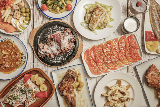 Overhead table of traditional Spanish dishes: sliced tomato salad, salted steak, cod with potatoes, roast lamb, stew, tuna salad and padron peppers on white wood