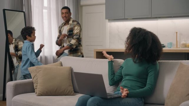 African American mother working online laptop on sofa while father and little boy child dancing distracting irritated woman businesswoman mom dad kid son family problems indoors home living room